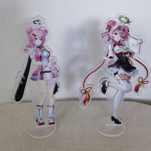Puede incluir: Dos figuras de acrílico transparente con personajes estilo anime de cabello rosa. La figura de la izquierda viste un conjunto rosa y blanco con una motosierra negra. La figura de la derecha viste un vestido negro y blanco con detalles rojos y sostiene un abanico.