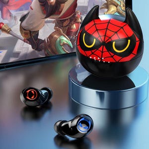 Op de afbeelding: Zwarte draadloze oordopjes met een oplaadcase. De case is zwart met een rood spinnenwebontwerp en gele ogen. De oordopjes hebben een cirkelvormig ontwerp met oranje en blauwe accenten. De oplaadcase staat op een zilveren standaard.