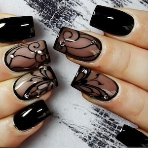 May include: Close-up of manicured nails. Some nails are painted solid black, while others feature a nude base with intricate black lace-like designs and small rhinestones. The nails are well-shaped and polished, showcasing a detailed nail art style.