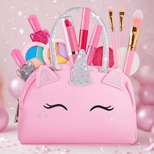 May include: A pink unicorn-shaped makeup bag filled with pink and silver nail polish, lipsticks, lip gloss, eye shadow, and makeup brushes. The bag has a silver horn, ears, and handle. The background is a soft pink.