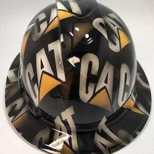 以下が含まれることがあります： 黒色の光沢仕上げのヘルメット。表面全体に白で繰り返される「CAT」の文字が特徴です。黄色とグレーの幾何学模様がデザインを引き立てています。ヘルメットには広いツバが付いています。