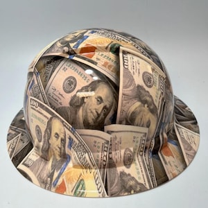 May include: A hard hat with a design featuring images of U.S. $100 bills. The hat is covered in the green and white bills, with the portrait of Benjamin Franklin visible. The brim and top of the hat are fully covered in the money design.