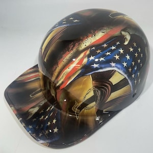 Op de afbeelding: Een glanzende zwarte veiligheidshelm met een patriottisch ontwerp met de Amerikaanse vlag, inclusief rode en witte strepen en witte sterren op een blauwe achtergrond. De helm heeft een gebogen klep.