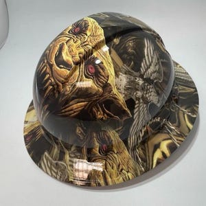 Puede incluir: Un casco de seguridad con un diseño detallado y texturizado que presenta una criatura con ojos rojos y una combinación de colores marrón y beige. El casco tiene un acabado brillante y un ala ancha.