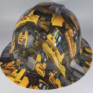 May include: A safety helmet with a repeating pattern of yellow construction vehicles on a black base. The hat has a brim and a shiny surface, suitable for construction or industrial work.