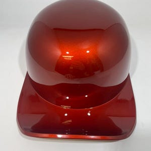 May include: A shiny, metallic red baseball helmet with a curved top and a visor. The helmet has a smooth, glossy finish and is designed to protect the head during sports activities. The helmet is shown against a white background.