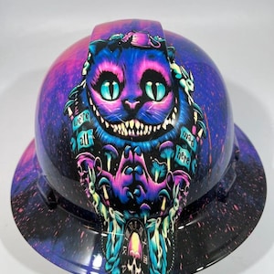 May include: A hard hat with a vibrant design featuring the Cheshire Cat from Alice in Wonderland. The cat is purple with a wide grin and teal eyes, surrounded by mushrooms and the text "we're all mad here."