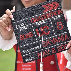 May include: Black graduation cap decorated with rhinestones. The cap features the words "GRADUATION," "DC," "SKYANIA," and "2025" in red. The date "MAY 2 2026" is also visible. The cap is held up by a person.