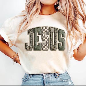 May include: Cream-colored t-shirt with the word "JESUS" in a distressed, textured design. The letters are outlined in a dark green color, with a checkered pattern on the "E" and a camouflage pattern on the "J", "S", and "U".