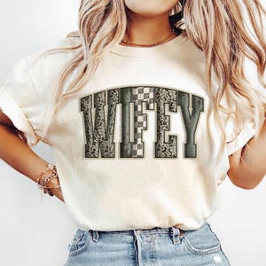 May include: Cream-colored t-shirt with the word "WIFEY" in a green and gray camouflage design. The letters have a collegiate style, with a checkered pattern on the "F" and a dark green outline. The shirt is made of a soft material.