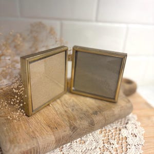 May include: A double-sided folding picture frame with a metallic gold-toned finish and thin, textured edges. The frame consists of two square glass panels connected by a central hinge, designed to hold two separate photographs side by side.