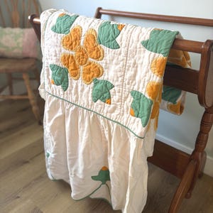 May include: A cream-colored quilt with a floral design in shades of yellow and green, draped over a wooden quilt rack. The quilt features a ruffled edge and a green border, showcasing a vintage aesthetic.
