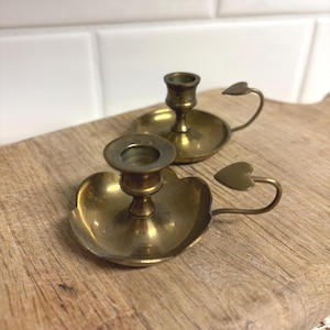 May include: Two vintage brass candle holders. Each holder has a round base with a heart-shaped handle. The candle holders are on a wooden surface. The candle holders are made of brass and have a warm, golden color.