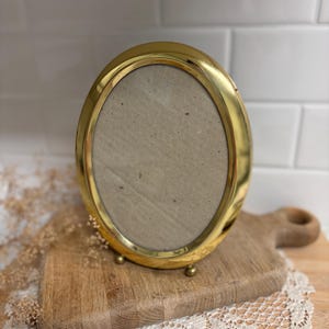 May include: An oval-shaped picture frame with a polished gold-tone metal finish. The frame features a smooth, rounded border and is supported by two small, spherical metal feet at the base. The center contains a plain, light beige cardboard insert.