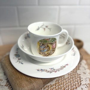 May include: A white ceramic teacup, saucer, and small bowl set. The teacup and saucer feature floral and Beatrix Potter illustrations. The set is on a wooden cutting board with a lace doily. The teacup has a handle.