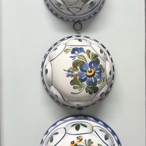 May include: A vertical wall hanging consisting of three white ceramic molds connected by metal rings. Each mold features a hand-painted floral design in pink, blue, or yellow with green leaves and blue trim along the scalloped edges.