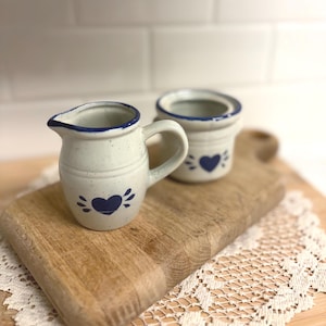May include: A small, off-white ceramic creamer and sugar bowl set with blue heart designs. The creamer has a handle and spout, while the sugar bowl is open. Both pieces have blue trim and are on a wooden cutting board.