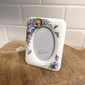 May include: A white ceramic picture frame with rounded corners, decorated with purple and green floral artwork. The frame has an oval opening for a photo and the text "Tomagafios" is printed on the frame.