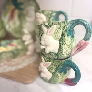 May include: Two stacked ceramic mugs with white rabbit relief designs, green leaf patterns, and teal handles. A matching plate and serving dish are visible in the background. The mugs have a carrot-shaped base.