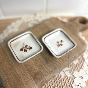 May include: Two small, square, ceramic dishes with a white background and brown floral designs. The dishes have brown rims and are set on a wooden cutting board. The dishes are likely for serving small portions.