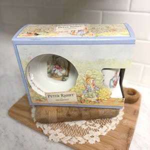 May include: A Peter Rabbit Wedgwood gift set in a light blue box. The set includes a small bowl and a cup, both featuring illustrations of Peter Rabbit. The box has a lid with a garden scene illustration.