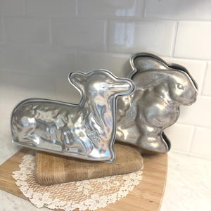 May include: Two vintage metal baking molds in the shapes of a lamb and a rabbit. The molds are silver-colored and sit on a wooden cutting board with a white lace doily. The lamb mold is approximately 7 inches long.