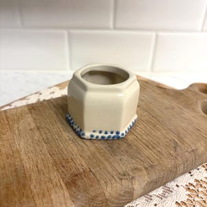 May include: A small, cream-colored ceramic planter with a hexagonal shape. The base features a blue dotted pattern. The planter sits on a wooden cutting board. The background is a white tiled wall.