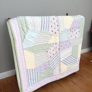 May include: A pastel patchwork quilt with a variety of fabrics in shades of green, yellow, blue, and purple. The quilt features striped and patterned sections, with embroidered star accents. The edges are finished with a lavender border.