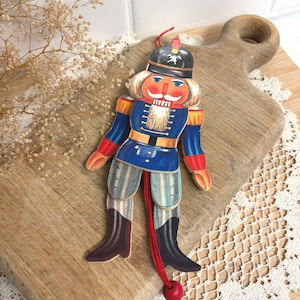 May include: A flat, articulated nutcracker ornament featuring a blue uniform with red and gold accents, a black hat with a white emblem, and a white beard. The figure has movable limbs connected by a red string with a small red bead at the bottom.