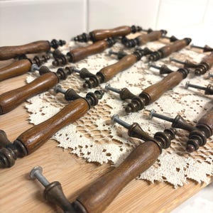 May include: A collection of vintage-style cabinet handles. Each handle features a dark brown, curved wooden grip with ornate metal hardware and a screw for mounting. The handles are arranged on a wooden surface.