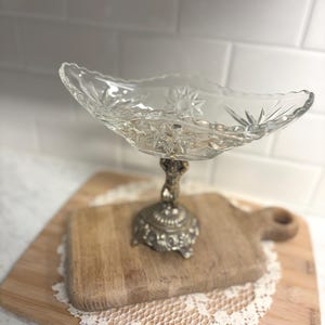 May include: A clear glass serving dish with a starburst pattern sits atop an ornate silver-colored metal base. The dish is displayed on a wooden cutting board with a decorative lace doily.
