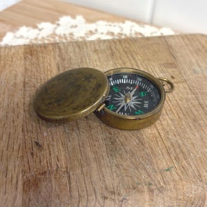May include: A vintage brass compass with an open lid, revealing a black and white dial with directional markings. The compass has a small loop for attachment and sits on a wooden surface. The compass is a navigational tool.