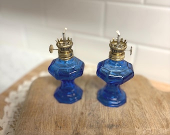 May include: Two antique cobalt blue glass oil lamps with brass accents. The lamps have a faceted design and are displayed on a wooden cutting board. The wicks are visible, suggesting they are ready for use. The lamps are a vintage decorative item.
