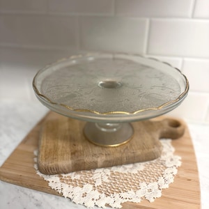 May include: A clear glass cake stand with a gold rim, resting on a wooden cutting board and a cream-colored lace doily. The stand has a decorative pattern on the plate.