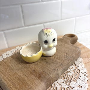 May include: A vintage ceramic chick figurine with a matching eggshell bowl. The chick is white with black eyes, a yellow beak, and a pink and yellow head. The eggshell is yellow with a white interior. The items are on a wooden cutting board.