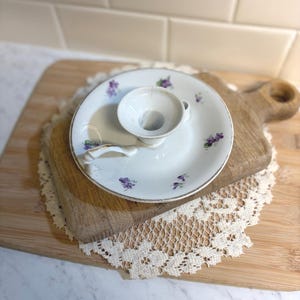 May include: A white porcelain candle holder with a saucer base, adorned with purple floral accents and a gold rim. The candle holder is placed on a wooden cutting board with a lace doily underneath, creating a vintage aesthetic.