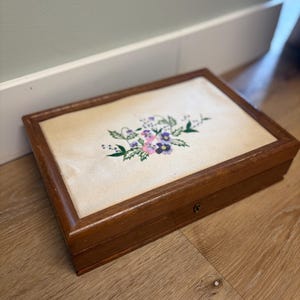 May include: A rectangular wooden box with a hinged lid featuring a central inset panel of off-white fabric. The fabric is decorated with an embroidered floral motif in shades of purple, pink, and green. A small metal keyhole is visible on the front.