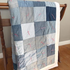 May include: A patchwork quilt with squares of denim in various shades of blue and gray. Pink thread accents the seams. The quilt has a white border with a dotted pattern and is draped over a wooden rack.