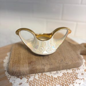 May include: A vintage, iridescent white ceramic bowl with gold trim and handles. The bowl features a decorative swirl design on the front and is set on a wooden cutting board.