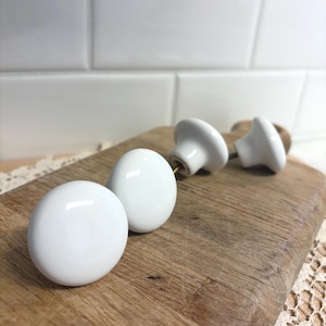 May include: A collection of white ceramic cabinet knobs with a glossy finish, arranged on a wooden cutting board. The knobs have a rounded shape and are designed for furniture. The background features white subway tile.