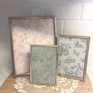 May include: Three vintage picture frames with ornate gold-colored borders. The frames display floral and toile patterns in pink, green, and blue. They rest on a wooden cutting board with a lace doily.