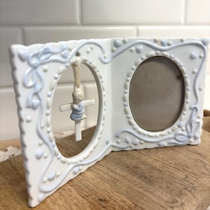 May include: White ceramic double photo frame with oval openings. The frame has a light blue floral and heart design. A small figurine of a child praying on a cross hangs in one opening. The frame is displayed on a wooden surface.