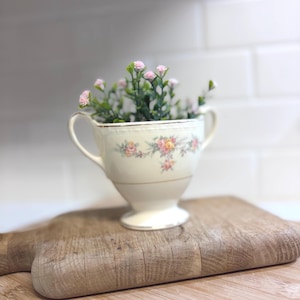 May include: A vintage teacup filled with small pink flowers and greenery. The teacup is cream-colored with a floral pattern in pink and yellow, and gold trim. It sits on a wooden cutting board, creating a charming, rustic display.