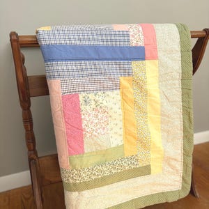 May include: A colorful patchwork quilt draped over a wooden chair. The quilt features squares and rectangles in various patterns and colors, including blue plaid, pink, yellow, and floral prints. The edges are finished with a green dotted fabric.