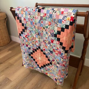 May include: A patchwork quilt featuring a multi-colored array of small fabric squares in various floral, geometric, and solid patterns. The design incorporates a central peach-colored diamond shape framed by black squares, creating a repeating pattern.