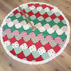 May include: A circular Christmas tree skirt featuring a patchwork design of red, green, and white fabric squares with various patterns. The skirt has a central opening, small green fabric ties between the squares, and a white eyelet lace trim.