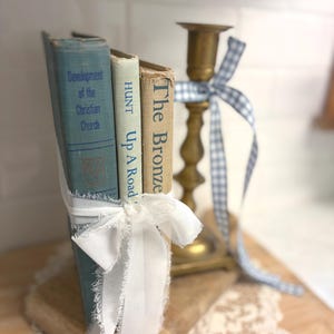May include: A vintage-style display featuring three antique books tied with a white ribbon, alongside a brass candlestick adorned with a blue and white gingham ribbon. The books have titles like "Development of the Christian Church".