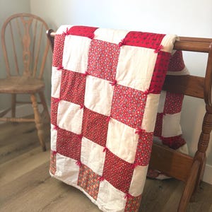 May include: A handmade quilt with a checkerboard pattern of red floral fabric and white squares. The quilt is draped over a wooden rack, showcasing its design. Pink yarn ties secure the squares. A wooden chair is in the background.