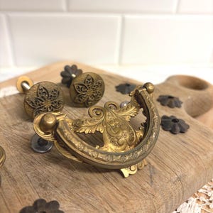 May include: Close-up of antique brass drawer pulls and knobs. The drawer pulls have an ornate, curved design with floral details. The knobs feature a floral pattern. Several dark metal decorative washers are also visible.