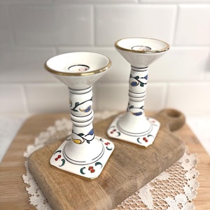 May include: Two white ceramic candle holders with gold rims and floral designs. The candle holders have a flared top and a decorative stem with blue, yellow, and green accents. They sit on a wooden cutting board.